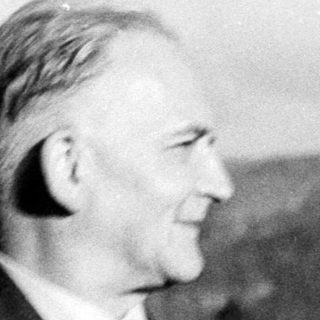 Man in suit stands outdoors, mountains visible in background.