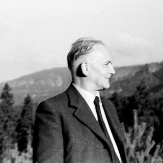 Man in suit stands outdoors, mountains visible in background.