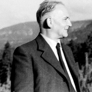 Man in suit stands outdoors, mountains visible in background.