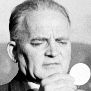Man in suit holds communion wafer outdoors, surrounded by trees.