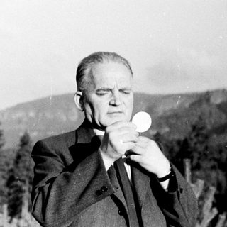 Man in suit holds communion wafer outdoors, surrounded by trees.