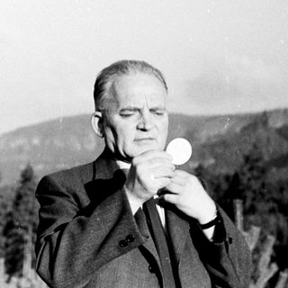 Man in suit holds communion wafer outdoors, surrounded by trees.