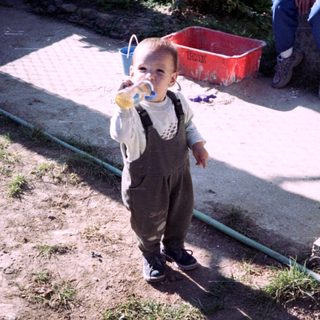 Baby in overalls stands drinking, adult sits nearby.