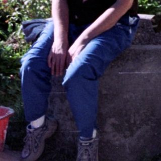 Baby in overalls stands drinking, adult sits nearby.