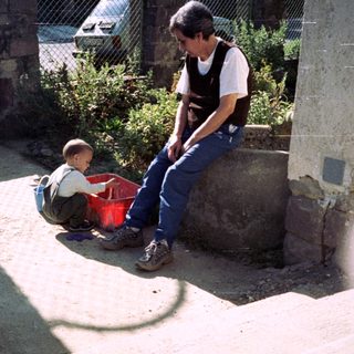 Man and child sitting outdoors, near a wall and greenery.