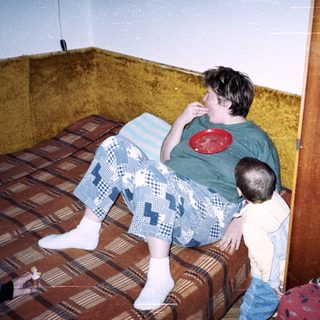 Here's a brief summary of the image: Boy sits on a bed, eating, with a toddler nearby.