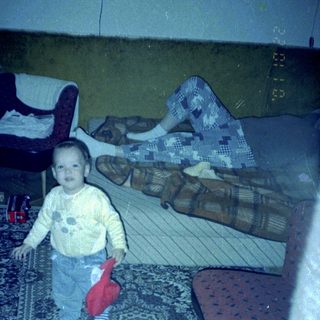 Child stands on rug, with person lying on couch.