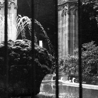 Fountain visible through bars, with a building and foliage.