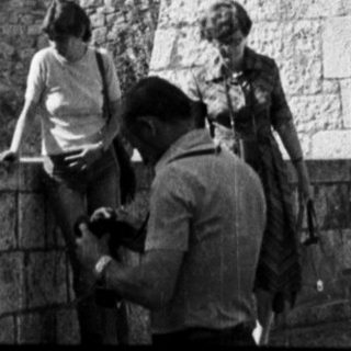 People near a stone building with a tower in background.