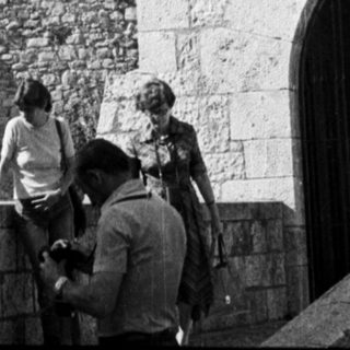 People near a stone building with a tower in background.