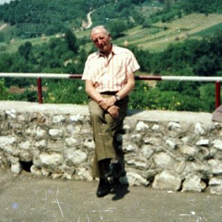 Elderly man seated near a stone wall overlooking a landscape.