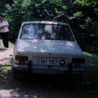 Man and woman stand near a vintage car in a wooded area.