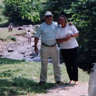 Man and woman stand near a vintage car in a wooded area.