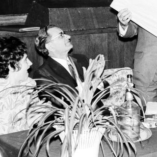 Men present at a table, holding papers and a large panel.