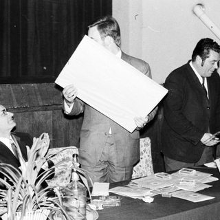 Men present at a table, holding papers and a large panel.