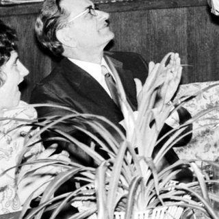 Men present at a table, holding papers and a large panel.