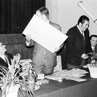 Men present at a table, holding papers and a large panel.