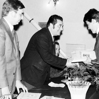 Two men handing a document to a third, formal setting.