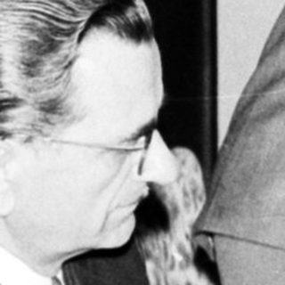 Two men in suits shake hands, appearing to exchange documents.