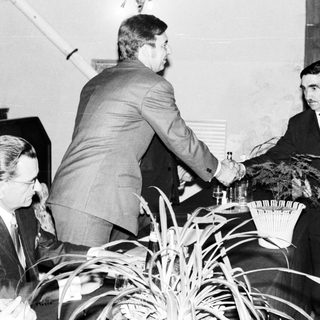 Two men in suits shake hands, appearing to exchange documents.