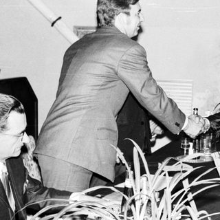 Two men in suits shake hands, appearing to exchange documents.
