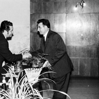 Men in suits shake hands near a table with plants.