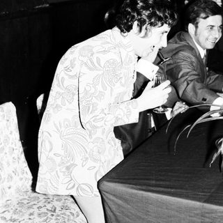 Woman in patterned dress leans over a table, men nearby.