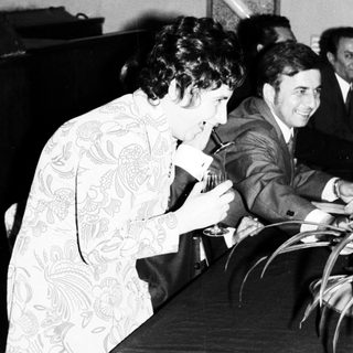 Woman in patterned dress leans over a table, men nearby.