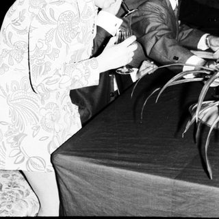 Woman in patterned dress leans over a table, men nearby.