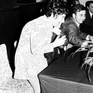 Woman in patterned dress leans over a table, men nearby.