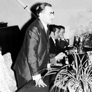 Man in suit stands near a table with people.