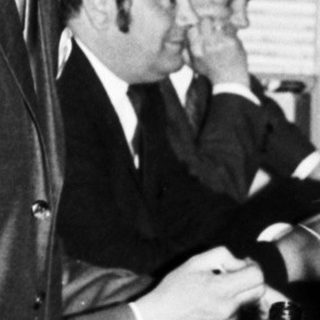 Man in suit stands near a table with people.