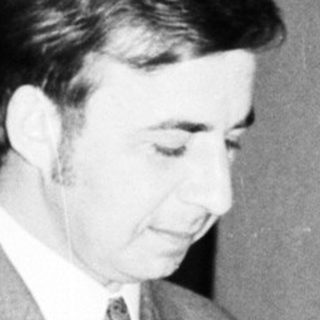 Man in suit stands near a framed document and table.