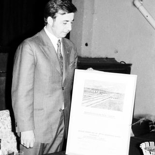Man in suit stands near a framed document and table.