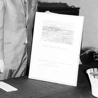 Man in suit stands near a framed document and table.