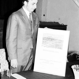 Man in suit stands near a framed document and table.