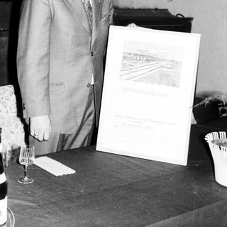 Man in suit stands near a framed document and table.