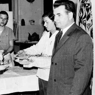 Here's a brief summary of the image: People serve food in a kitchen setting.