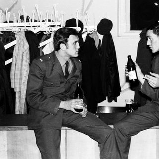 Two men in military uniforms sit, holding bottles.