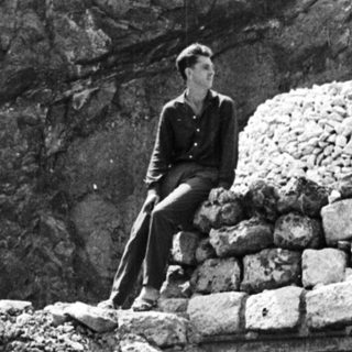 Man sits atop a stone structure near a rock face.