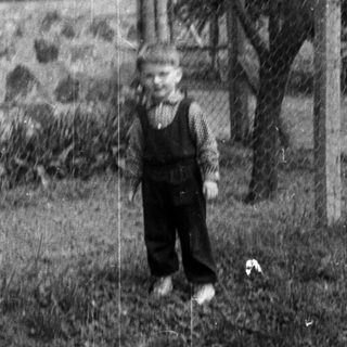 Here's a brief summary of the image: Young boy stands in a yard near a stone building.
