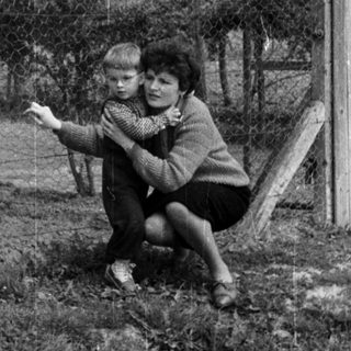 A woman holds a child outdoors in a grassy area.