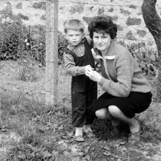 A child and woman stand near a fence and building.