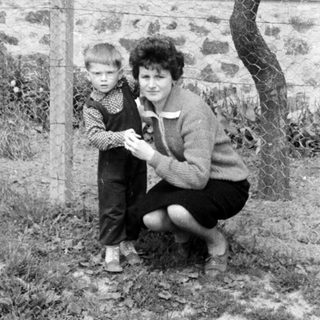 A child and woman stand near a fence and building.