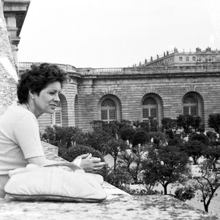 Woman sits on a balcony overlooking an elegant building.