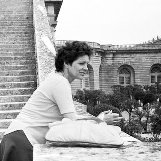 Woman sits on a balcony overlooking an elegant building.