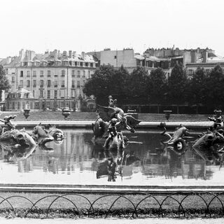 Fountain with statues and city buildings reflected in water.