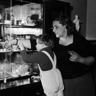 Child reaching for items in a cabinet, woman watching.