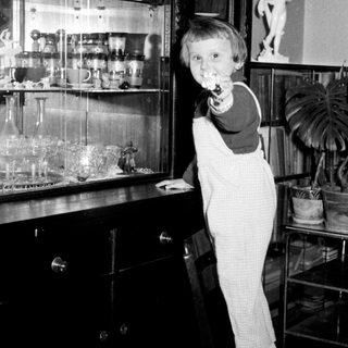 Boy in overalls stands on a chair near a cabinet.