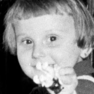 Boy in overalls stands on a chair near a cabinet.